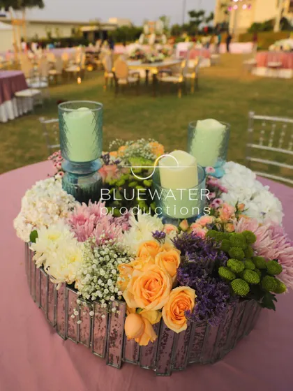 A beautiful tabletop centerpiece for an outdoor wedding, featuring a mix of pastel florals and candles in textured glass holders.