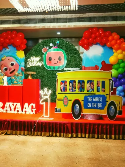 A vibrant Cocomelon themed stage decoration for a first birthday party. This setup includes a large character backdrop, a custom Cocomelon school bus prop, and colorful balloon arches, accurately bringing the popular show to life.