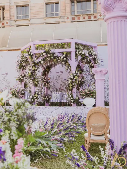 A beautiful pastel purple gazebo-style mandap, adorned with lush floral garlands. The printed backdrop and matching cushions create a cohesive and charming English garden theme.