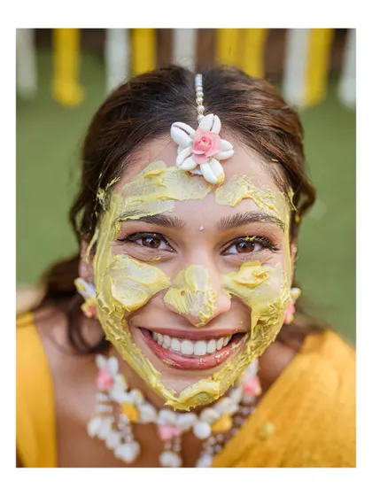 A beautiful close-up of the bride, her face adorned with turmeric and a big, bright smile. Her floral jewelry adds a delicate touch.