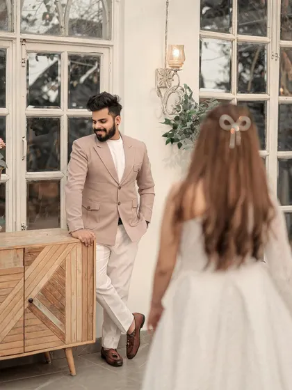 A candid, story-driven shot. The groom waits with a smile as his bride approaches, creating a sense of anticipation and romance in this beautifully lit indoor setting.