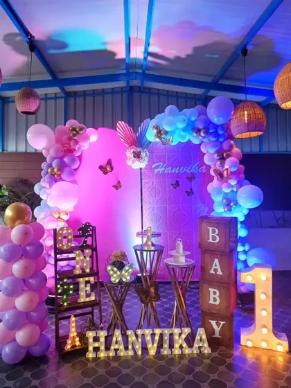 A whimsical butterfly-themed setup for Hanvika's first birthday. The decor uses dramatic pink and purple lighting to create a magical, glowing effect.