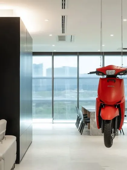 A front view of the suspended red scooter, framed against the backdrop of the city skyline visible through large windows. This placement connects the company's innovative product directly with the urban environment it is designed for.