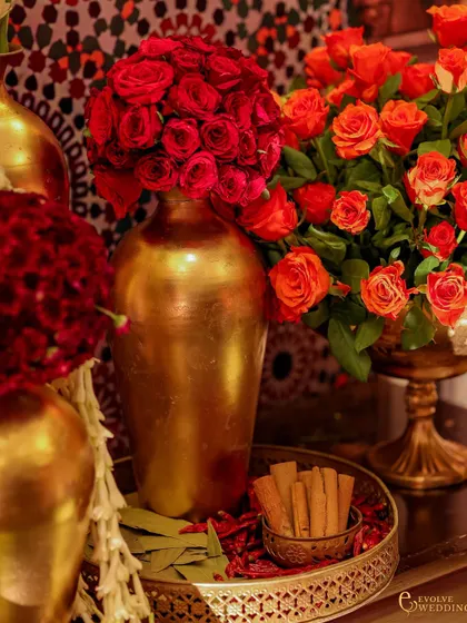 A detail shot of golden vases filled with red and orange roses, with a tray of cinnamon sticks in the foreground. The image captures the warmth and richness of the decor.