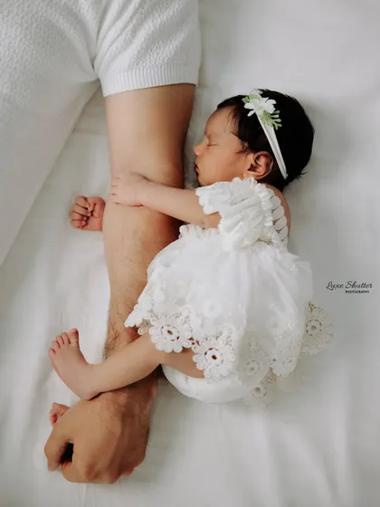 A tiny newborn girl in a delicate lace dress, sleeping peacefully while holding onto her father's arm. A creative and heartwarming shot.
