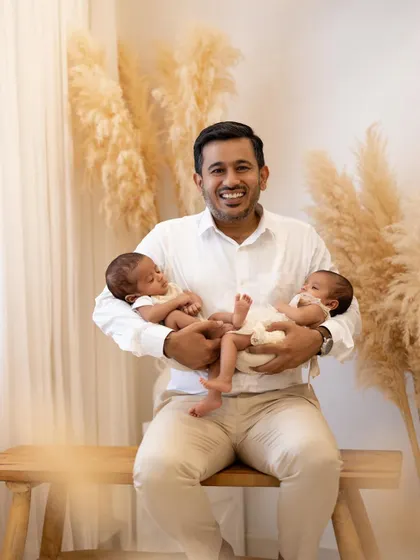 A proud father's smile says it all. He sits on a wooden bench in the studio, holding both of his newborn twins securely in his arms, beaming with happiness.