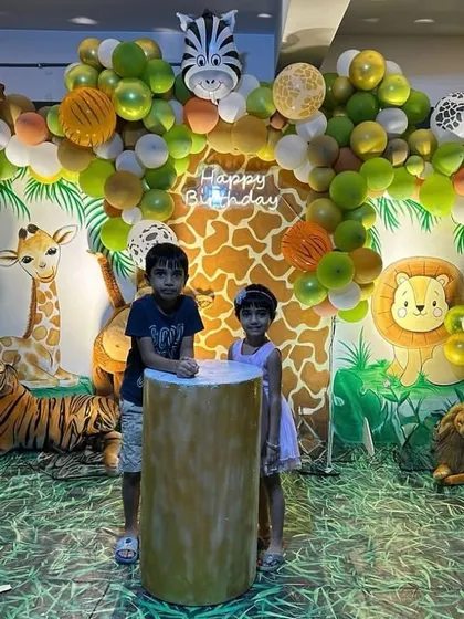 A vibrant jungle-themed birthday backdrop with kids posing for a photo. The setup includes a detailed balloon arch with animal prints and a floor covered with artificial grass matting.