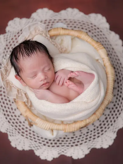 A top-down view of a baby swaddled in a cream-colored wrap, nestled in a wicker basket on a lace doily.