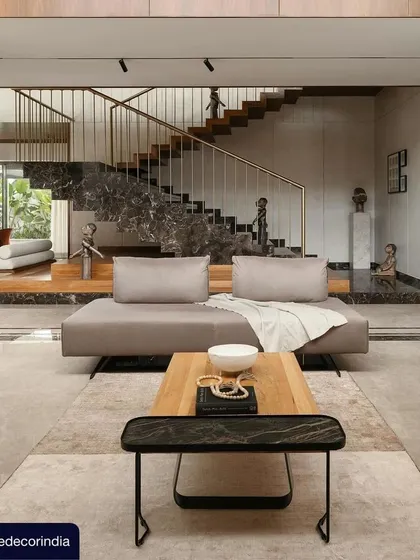 This image from the Elle Decor feature showcases the grand living and circulation space in Villa Shakthi. The composition highlights the interplay of materials, from the dark marble of the staircase plinth to the warm wood and brass accents of the railing.