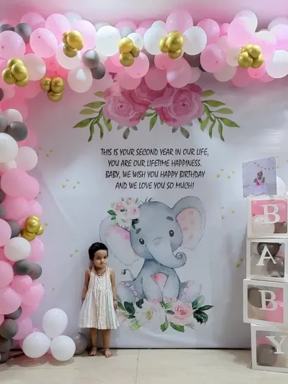 An adorable elephant-themed birthday party. The large, personalized backdrop is framed by a pink, white, and grey balloon arch, with "BABY" blocks on the side.