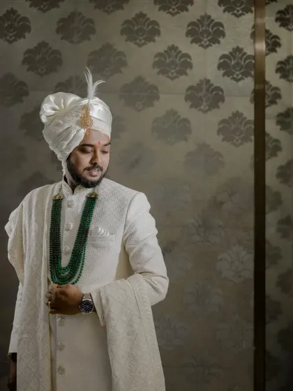A portrait of the groom in a classic white sherwani and an emerald green necklace, exuding elegance.