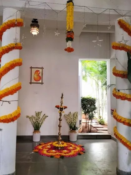 The entrance to a home decorated for a traditional ceremony. Pillars are wrapped in marigold garlands, and a floral rangoli with a brass lamp creates a warm and auspicious welcome.
