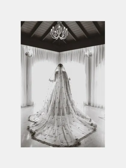 A dramatic black and white photo of a bride from behind, her long, elaborate veil filling the frame as she stands before a bright window.