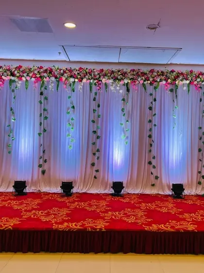 A clean and minimalist stage backdrop perfect for any event. It features a full wall of white curtains with hanging green vines and a floral border at the top, all beautifully illuminated by spotlights from the floor.