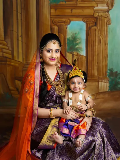 A regal portrait of a mother and her baby Krishna. The rich colors of their sarees and the traditional backdrop create a stunning and majestic photograph.