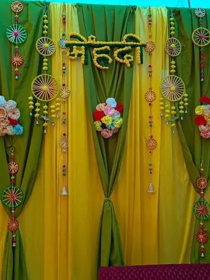 A beautifully draped Mehendi backdrop using yellow and green fabric, accented with small floral bouquets and wheel-shaped hangings.