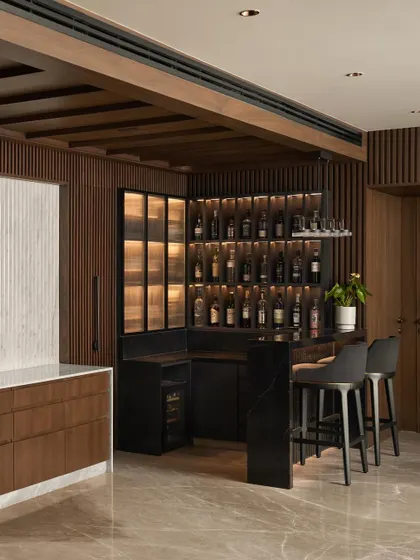 The bar area is seamlessly integrated with the entrance, featuring a backlit display for spirits, a wine fridge, and a black marble countertop. Fluted wood paneling connects the bar to the rest of the living space.