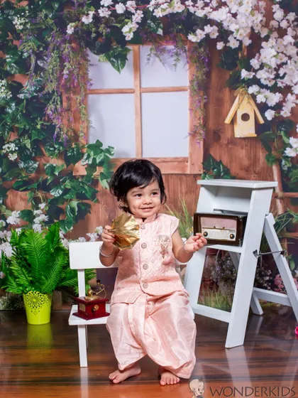 A handsome little boy in a traditional pink kurta and dhoti, smiling brightly in a garden-themed studio setup. His outfit and the floral backdrop create a perfect picture for any celebration.