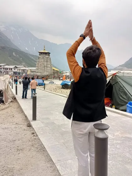 A devotee stands with hands joined in prayer, looking towards the Kedarnath temple. This image captures a personal moment of faith and surrender against the vast Himalayan backdrop.