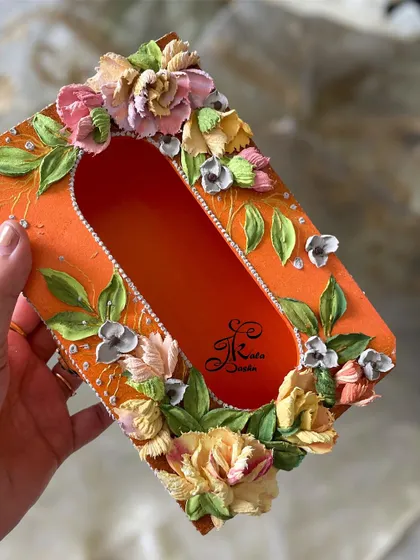 A vibrant orange tissue box adorned with sculpted flowers in soft, buttery shades of yellow and pink. The pearl details add a touch of elegance to this bright and sunny piece.