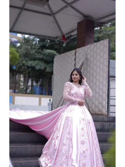 A model wearing a beautiful pink ball gown with a long, flowing train. The gown features delicate embroidery and sheer sleeves, creating a romantic and fairytale-like look for a reception or pre-wedding shoot.