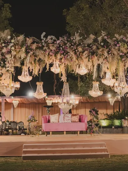 A stunning reception stage featuring a massive arch of pastel flowers and pampas grass, with hanging chandeliers and a plush pink sofa for the couple.