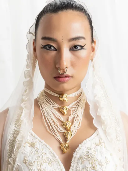A direct and striking portrait focusing on a multi-strand pearl choker with gold motifs. The styling is clean and minimalist, with a sharp winged eyeliner and nose ring adding a touch of edge to the otherwise delicate jewelry.