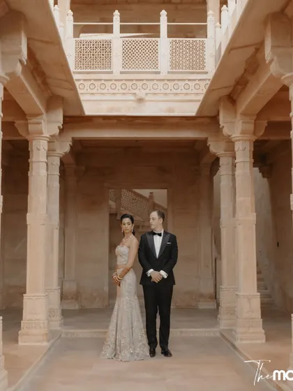 A formal portrait set within the grand, pillared hall of Suryagarh. The symmetrical composition and the couple's poised stance create an image of timeless elegance and sophistication.