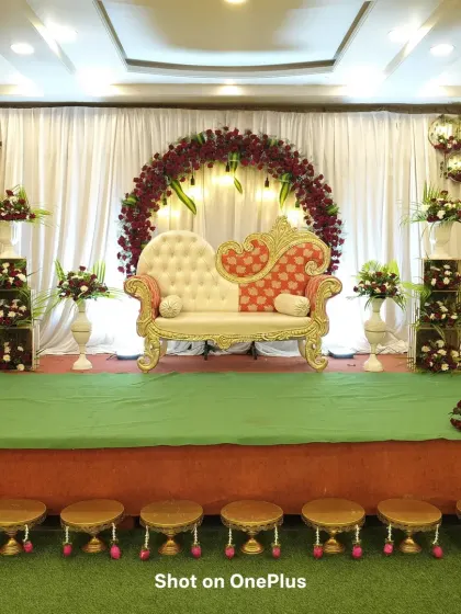 A stage prepared for a traditional function, featuring a circular arch of red roses against a white curtain. The ornate two-toned sofa is the centerpiece, ready for the main event.