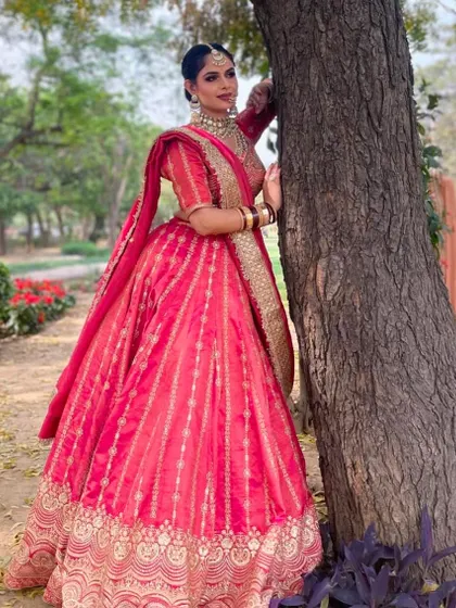 A full-length shot of a bride in a vibrant pinkish-red lehenga, perfect for an outdoor day wedding. The color stands out beautifully against the natural background.