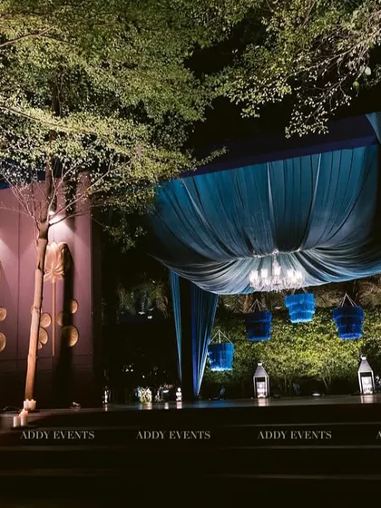 An atmospheric shot of the blue draped lounge, nestled among trees. The uplighting on the trees and the warm glow of the candles create a magical, moody environment for an evening sangeet or cocktail party.