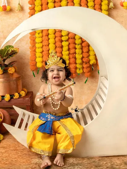A playful and expressive shot of a baby dressed as Krishna, shouting with joy on the moon prop.