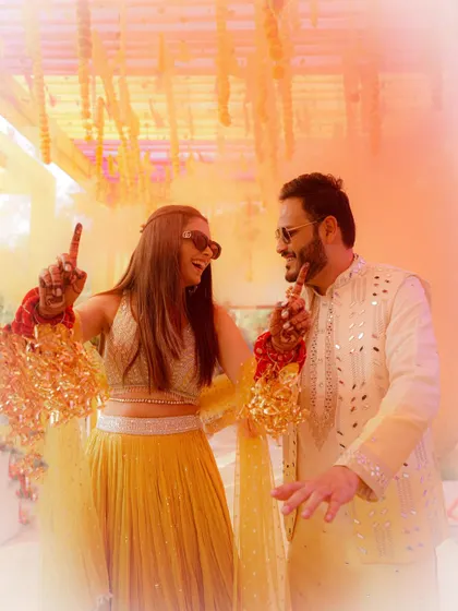 A fun, high-energy shot of the couple dancing with sunglasses on, the colorful background adding to the celebratory vibe of their Haldi.