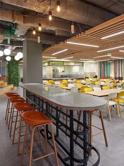 A cafeteria with an industrial vibe, featuring an exposed ceiling, concrete-topped high table, and a mix of seating types. The green wall in the background adds a touch of nature.