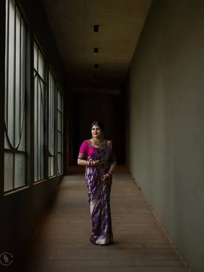 A full-length portrait in a dimly lit corridor, using the architecture to create a dramatic and elegant frame for the bride-to-be in her beautiful purple saree.