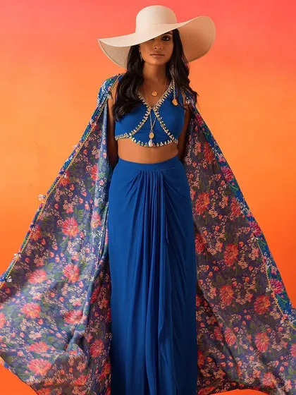 A perfect destination wedding look. This ensemble features a royal blue draped skirt and bralette, paired with a dramatic floral-printed cape and a wide-brimmed hat.