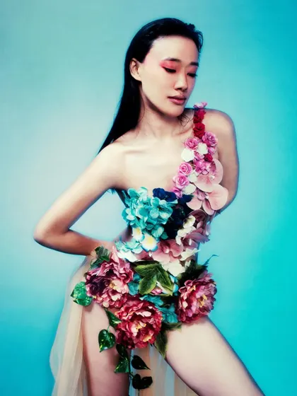 A close-up shot of the model in the floral bodysuit against a bright blue background. Her serene expression and the vibrant colors create a striking contrast, blending beauty with sorrow.