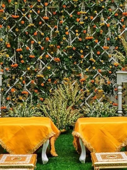 A beautiful haldi seating area featuring a floral wall of marigolds and greenery against a white lattice. The low, traditional chowkis covered in yellow fabric are perfect for the ceremony rituals.