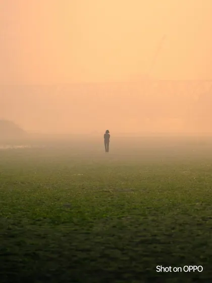 A lone figure stands in a vast, grassy field, enveloped in the thick morning fog with a bridge faint in the background. The image conveys a sense of solitude and mystery.