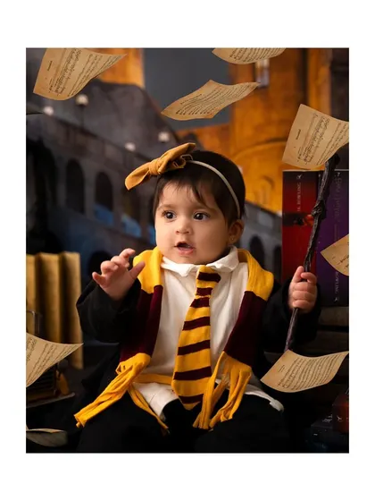 A baby girl dressed as a little witch from Harry Potter, complete with a Gryffindor scarf and a magic wand.