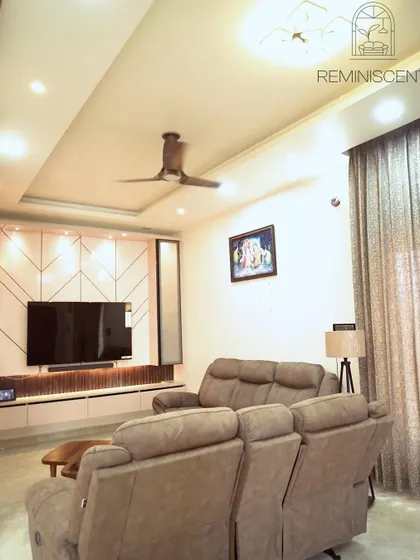 A wider view of the living room, showing the stylish TV unit, comfortable recliner sofa, and the elegant false ceiling with cove lighting.