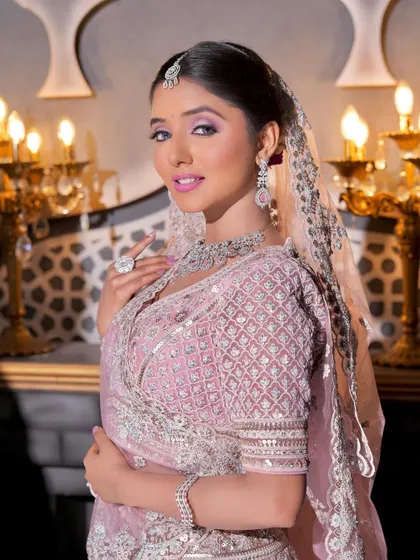 A beautiful bride looking gorgeous for her wedding in a regal pink lehenga. The makeup is glamorous, with a focus on dramatic eyes and luminous skin, set against an opulent backdrop.