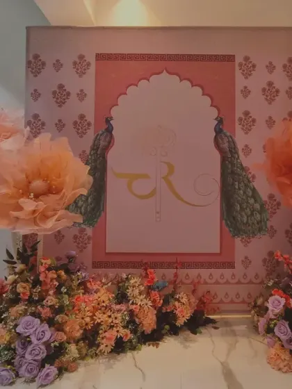 A traditional Indian-themed backdrop perfect for a sangeet or mehendi. It features a Rajasthani arch design with peacock motifs and is decorated with large, colorful paper flowers.
