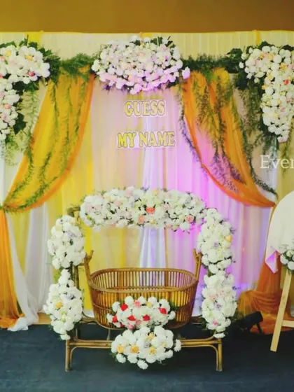 A beautiful setup for a prince's naming ceremony. The decor includes a flower-adorned cradle, a name reveal stand, and a backdrop of white and yellow drapes with lush floral arrangements.