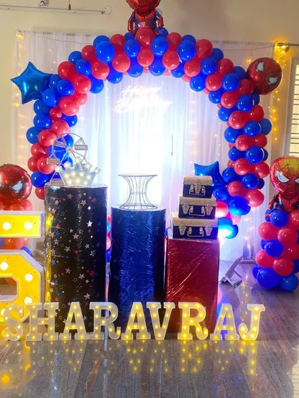 A dynamic Spiderman-themed birthday party for a 5th birthday. The setup includes a red and blue balloon arch, character foil balloons, and light-up marquee letters and numbers, all arranged in a home living room.