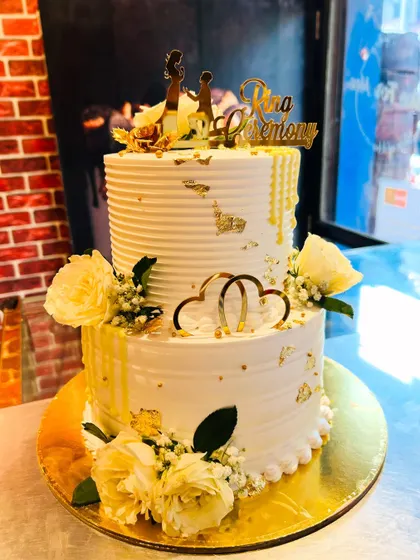 An elegant two-tier Roka ceremony cake in white and gold. It's decorated with fresh white roses, baby's breath, and gold leaf for a touch of luxury.