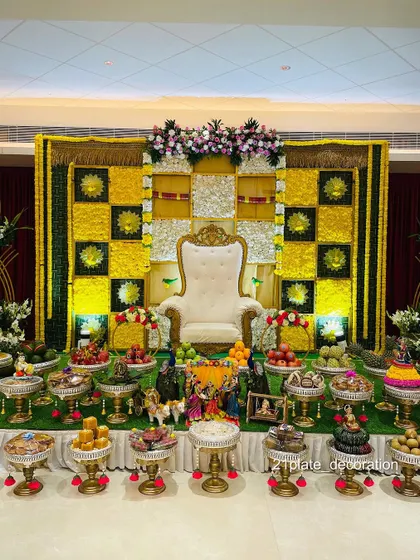 A grand and traditional baby shower stage with a yellow and green checkered floral backdrop. The extensive display of decorated plates and props makes it a complete and auspicious setup.