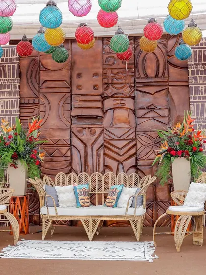 A cozy seating nook with peacock-style wicker furniture set against a wall of carved wooden panels. The colorful hanging lanterns add a playful and festive touch.