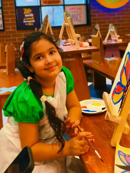 A young artist smiles proudly next to her butterfly painting. Our workshops provide all the materials and a supportive environment to let creativity flourish.