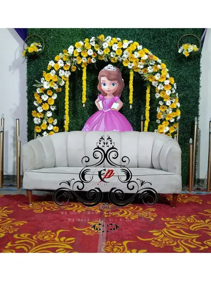 A sweet and simple backdrop for a half-saree ceremony. The setup includes a green grass wall, a floral arch, and a cutout of Princess Sofia.